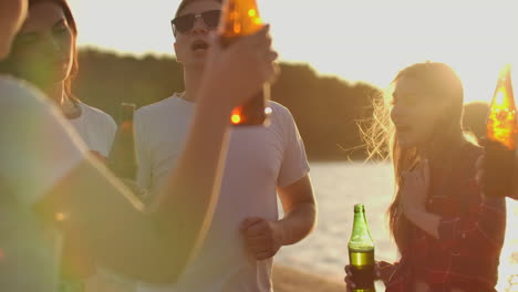 Group-of-students-celebrate-the-end-of-the-semester-with-beer-and-pop-music-on-the-beach.-They-are-dancing-on-the-open-air-party-at-sunset-in-hot-summer-evening.